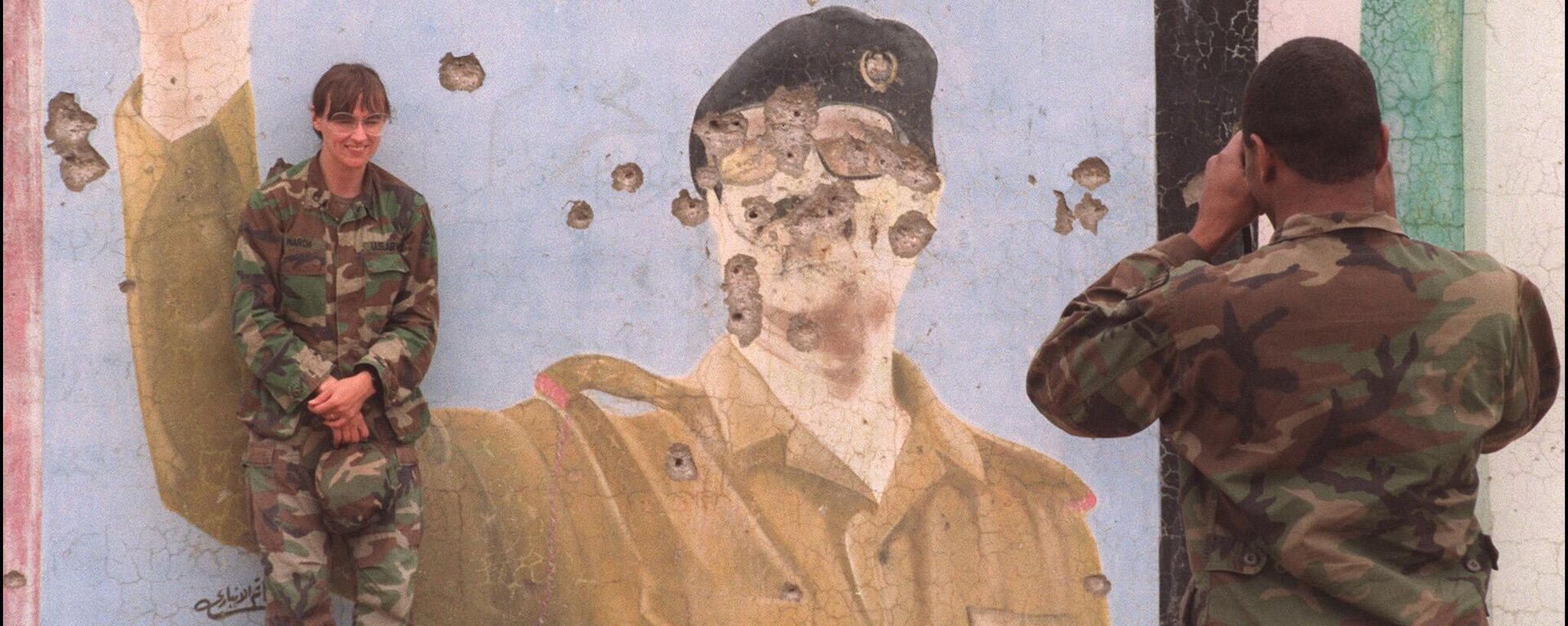 01 Mayıs 1991'de  bir ABD askeri,  meslektaşının Irak Devlet Başkanı Saddam Hüseyin'in kurşunlanmış devasa bir grafiti portresinin önünde poz verirken fotoğrafını çekiyor  - Sputnik Türkiye, 1920, 27.06.2023