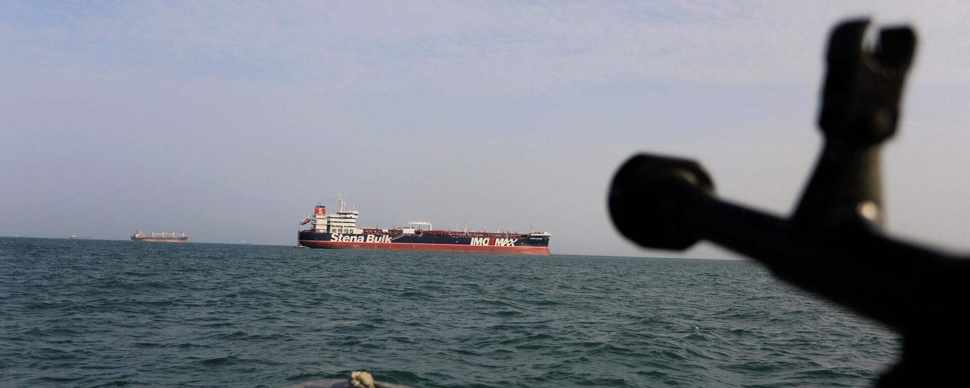In this Sunday, July 21, 2019 photo, a speedboat of Iran's Revolutionary Guard trains a weapon toward the British-flagged oil tanker Stena Impero, which was seized in the Strait of Hormuz on Friday by the Guard, in the Iranian port of Bandar Abbas. Global stock markets were subdued Monday while the price of oil climbed as tensions in the Persian Gulf escalated after Iran's seizure of a British oil tanker on Friday.  - Sputnik Türkiye, 1920, 06.05.2026