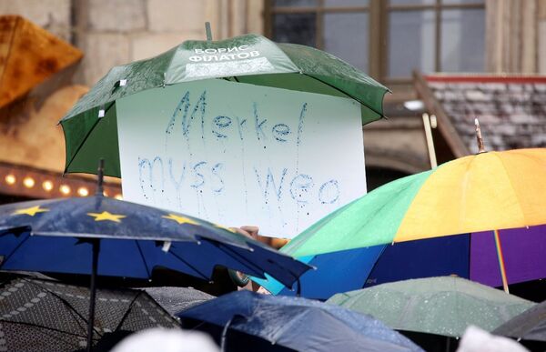 Dresden'de yoğun yağmur yağışı nedeniyle protesto gösterisine şemsiyelerle gelen eylemcilerden biri, şemsiyesinin altında 'Merkel gitmeli' yazılı bir pankart taşıdı. - Sputnik Türkiye